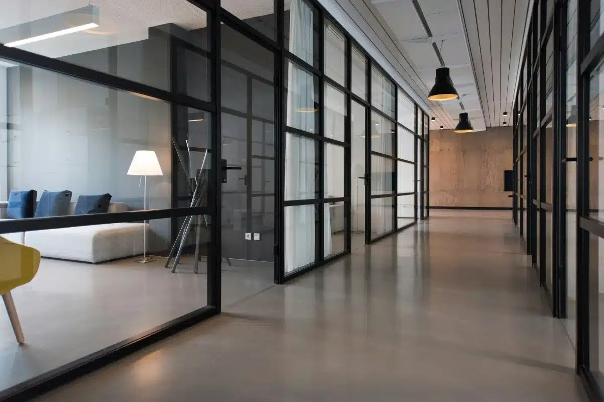 commercial-cleaning-img-1 Long glass-walled office corridor with black metal frames and a seating area to the left, including a white sofa and floor lamp.