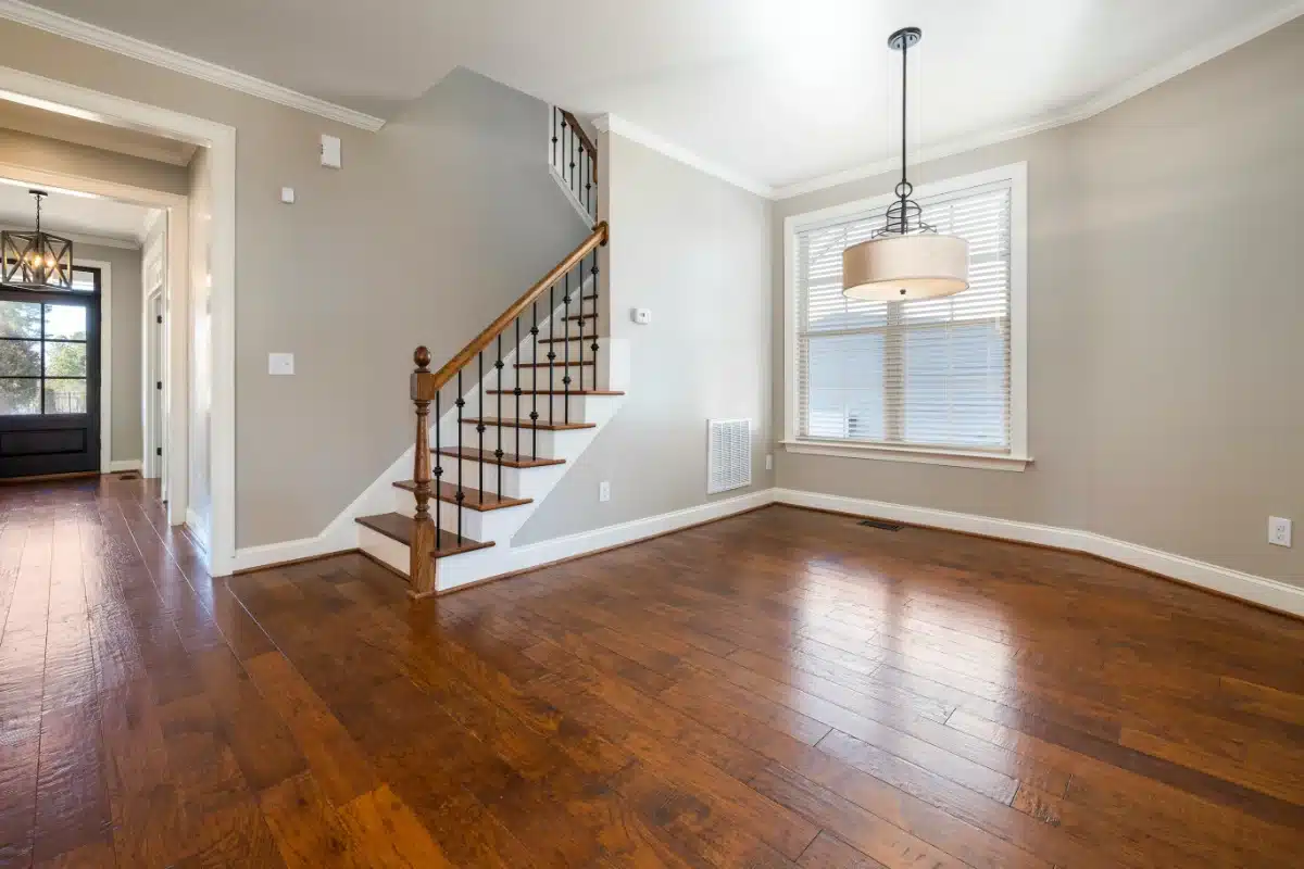 real-estate-cleaning-img-1 Bright empty living room with hardwood floors, a staircase with wooden railing, and a large window with blinds; contemporary chandelier hangs from the ceiling.